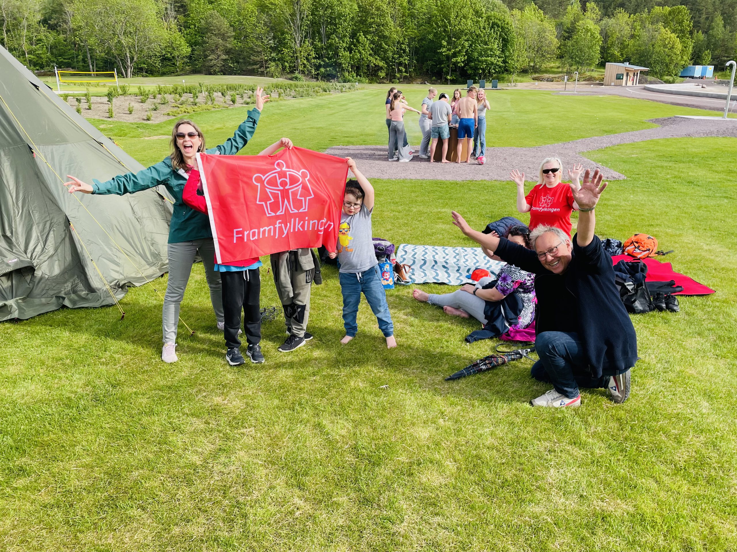 Barn og voksne, sitter på huk eller står ved siden av et telt på en gresslette. De vinker til fotografen og ler, viser fram et flagg med logoen til Framfylkingen på. Rødt flagg med tegning i hvitt av fire barn i ring som holder hender.